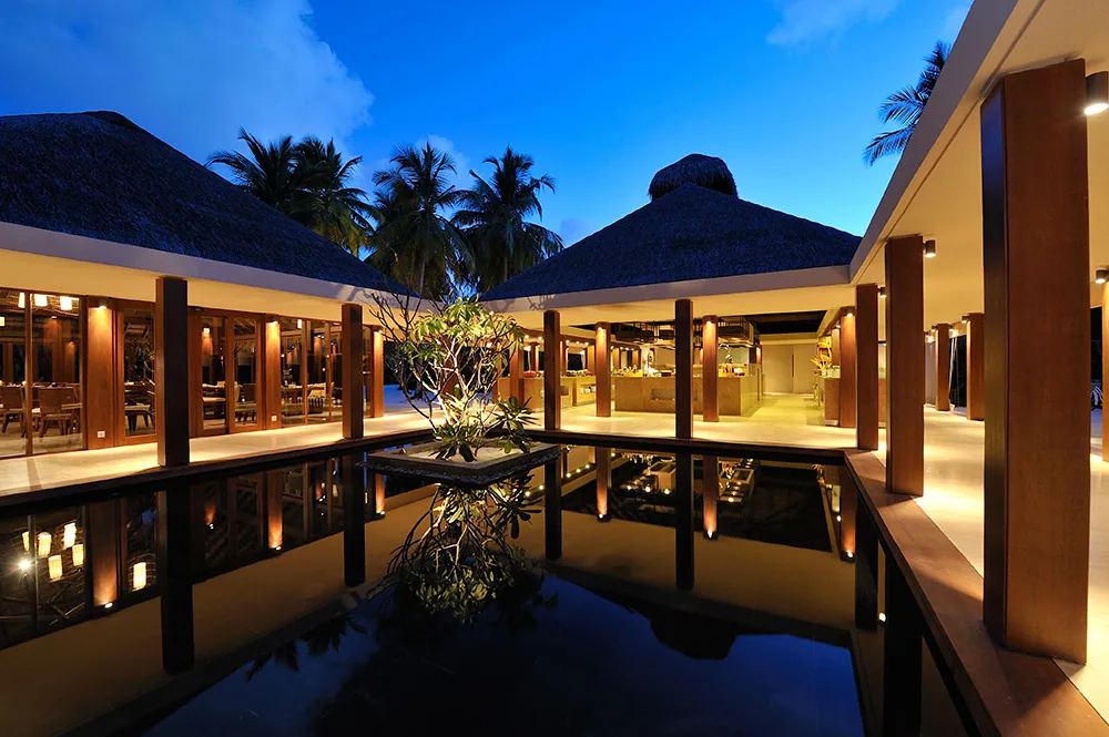 Ayada Maldives, dining, Gaafu Dhaalu Atoll, Maldives, Magu main restaurant pavilion at dusk: twin thatched-pyramid roofs flanking a central reflecting pool with frangipani tree planted at the axis, columned open walkways carrying the international-buffet themed-night programme.
