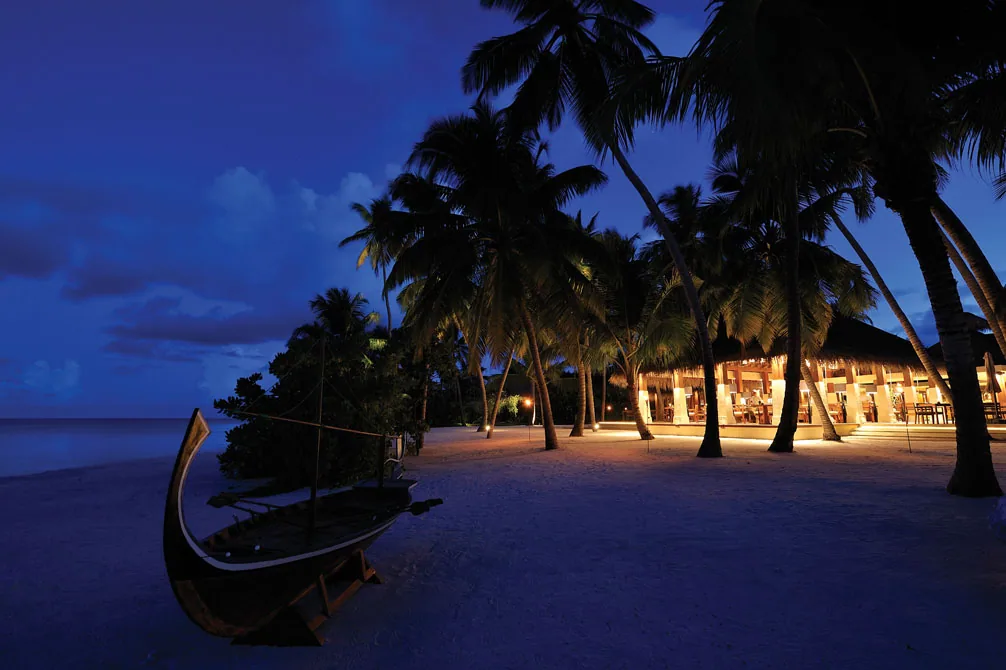 Ayada Maldives, dining, Gaafu Dhaalu Atoll, Maldives, Magu pavilion seen from the beach at blue hour, traditional Maldivian dhoni hull beached on white sand, palm canopy overhead, lit interior glowing through the open-side dining hall.