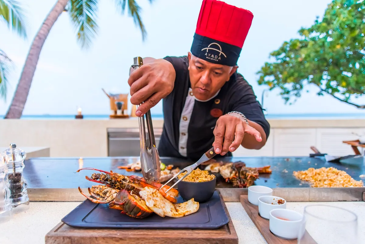 Ayada Maldives, dining, Gaafu Dhaalu Atoll, Maldives, Mizu teppanyaki chef in red toque and black AYADA-branded uniform, plating spiny lobster and scallop on the slate board at the outdoor counter, ocean horizon behind, ten-seat venue under the Maldivian night sky.