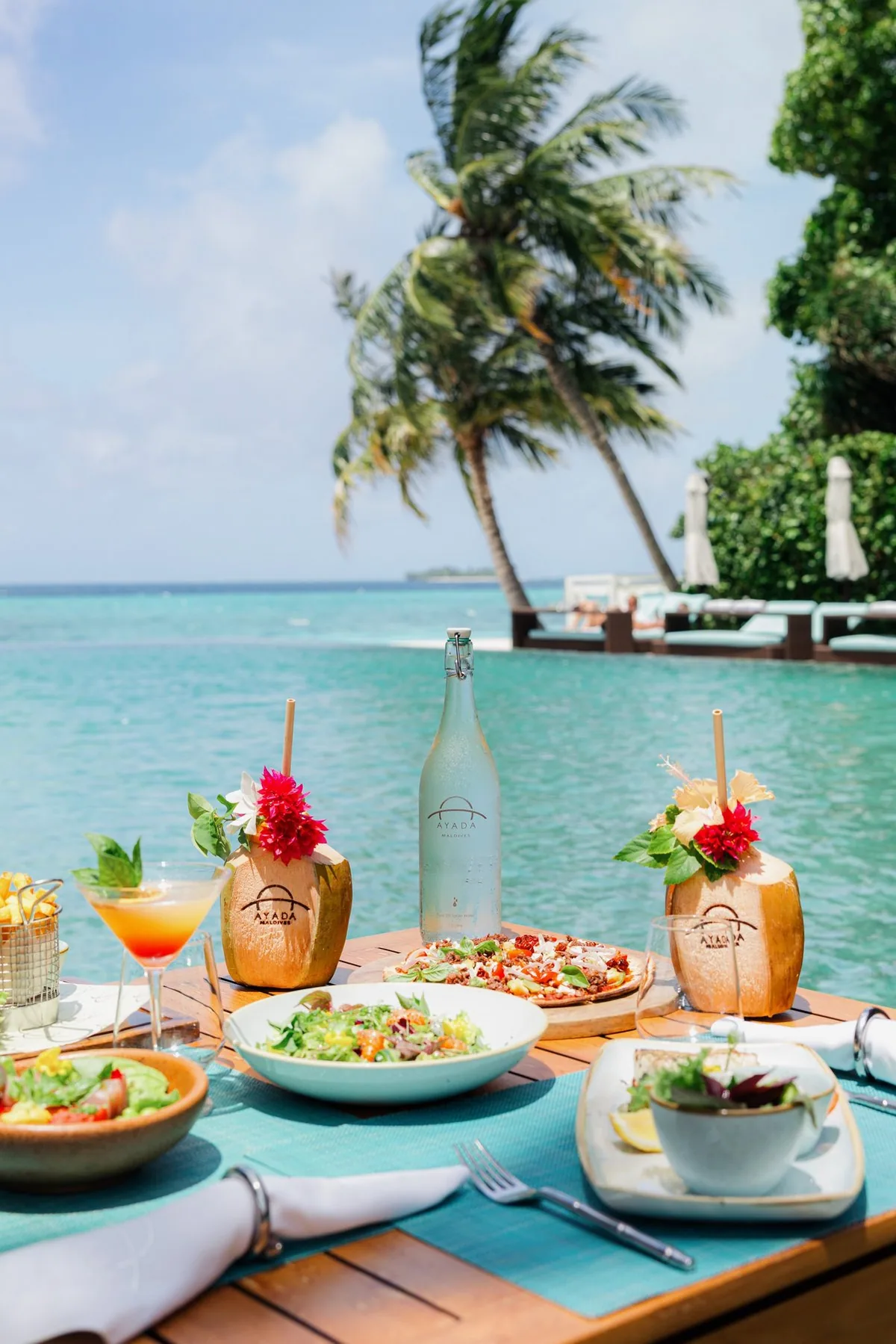 Ayada Maldives, dining, Gaafu Dhaalu Atoll, Maldives, Zero Degree poolside lunch on a turquoise woven runner, AYADA-branded coconut-shell tiki cocktails with hibiscus garnishes, branded glass bottle centrepiece, pizza and Mediterranean small plates, infinity pool edge stepping into the lagoon beyond.