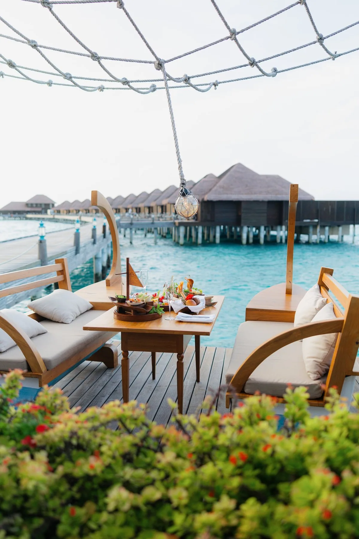 Ayada Maldives, dining, Gaafu Dhaalu Atoll, Maldives, Île de Joie wine and cheese island: an intimate two-seat setting on a small overwater deck, rope-net pergola overhead with hand-blown glass float lantern, Maldivian dhoni-prow chair backrests, overwater villa cluster on the jetty bridge as backdrop.
