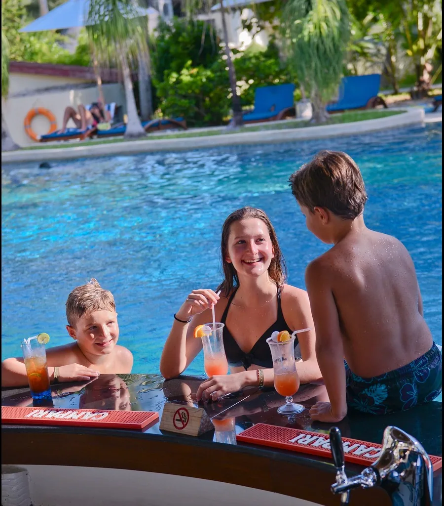 Bandos Maldives, bar, North Malé Atoll, Maldives, Pool swim-up bar: a family group of three young guests at the swim-up bar counter with cocktail drinks, the Campari-branded bar mat visible, sun loungers across the pool with another swimmer, dense coconut palm canopy beyond, the property's all-ages family-oriented bar identity captured in single frame.