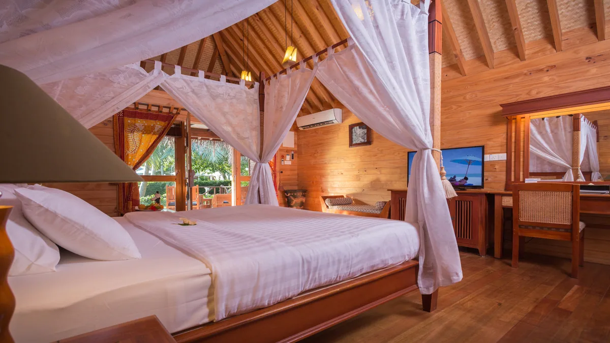 Bandos Maldives, villa beach, North Malé Atoll, Maldives, Garden Villa interior: distinctive vaulted timber-rafter ceiling with all-timber wall paneling (signature traditional Maldivian villa vocabulary), king bed with white mosquito-net canopy, dressing table with rattan-cane chair, dark walnut floor, open glass doors revealing the tropical garden setting.