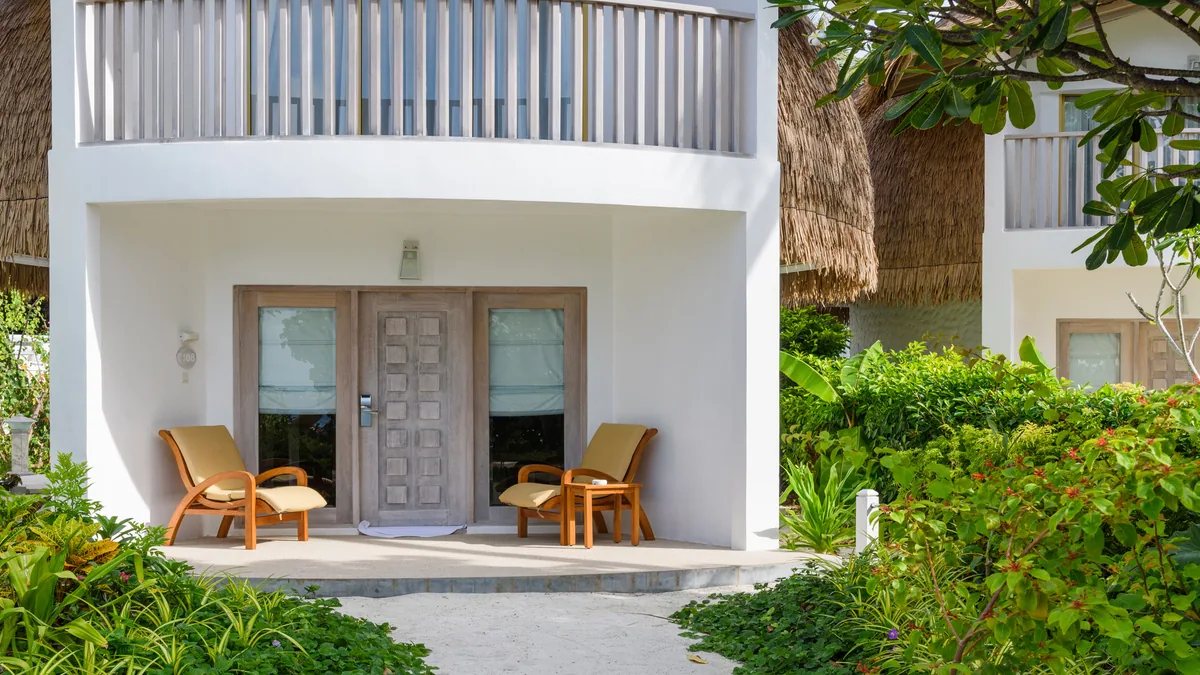 Bandos Maldives, villa beach, North Malé Atoll, Maldives, Premium Beach Villa exterior: distinctive two-storey configuration with white curved-stucco walls and traditional thatched roof at the top, ground-floor entry flanked by twin timber armchairs, an upper-floor balcony with cream railing, the sandy path approach lined with tropical greenery.