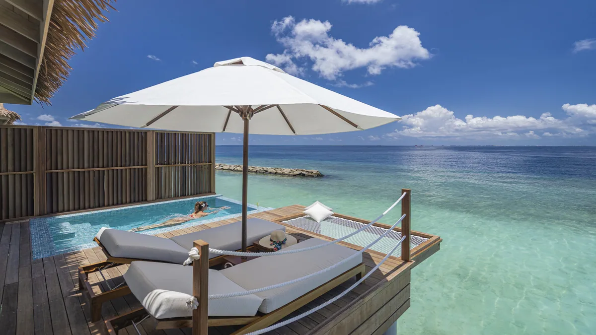 Bandos Maldives, villa overwater, North Malé Atoll, Maldives, Sunset Water Villa private deck: twin grey-cushioned sun loungers on the timber deck under a large white parasol, an infinity-edge plunge pool with a guest visible swimming, the over-water hammock-net stretched at the right edge for relaxation, slatted timber privacy fence, the breakwater-rock detail in the lagoon visible at the mid-distance, ocean horizon.