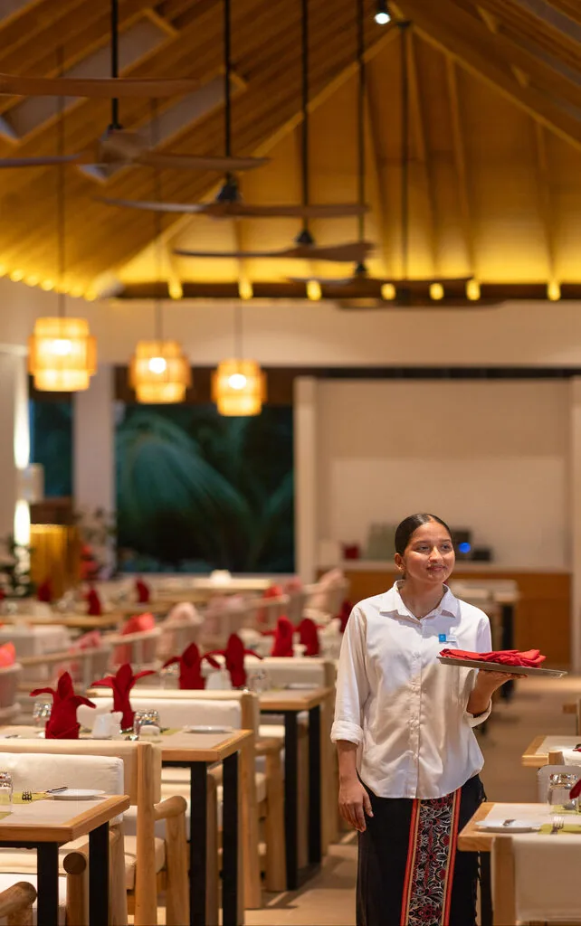 Bandos Maldives, dining, North Malé Atoll, Maldives, Principal buffet restaurant interior: distinctive golden-bamboo woven cathedral ceiling with a row of teardrop ceiling fans, line of orange pendant lanterns, blonde-wood dining chairs with white seat cushions, the tables set with red napkin pyramid presentations, a server in white shirt and patterned wrap-skirt holding a tray in mid-frame.