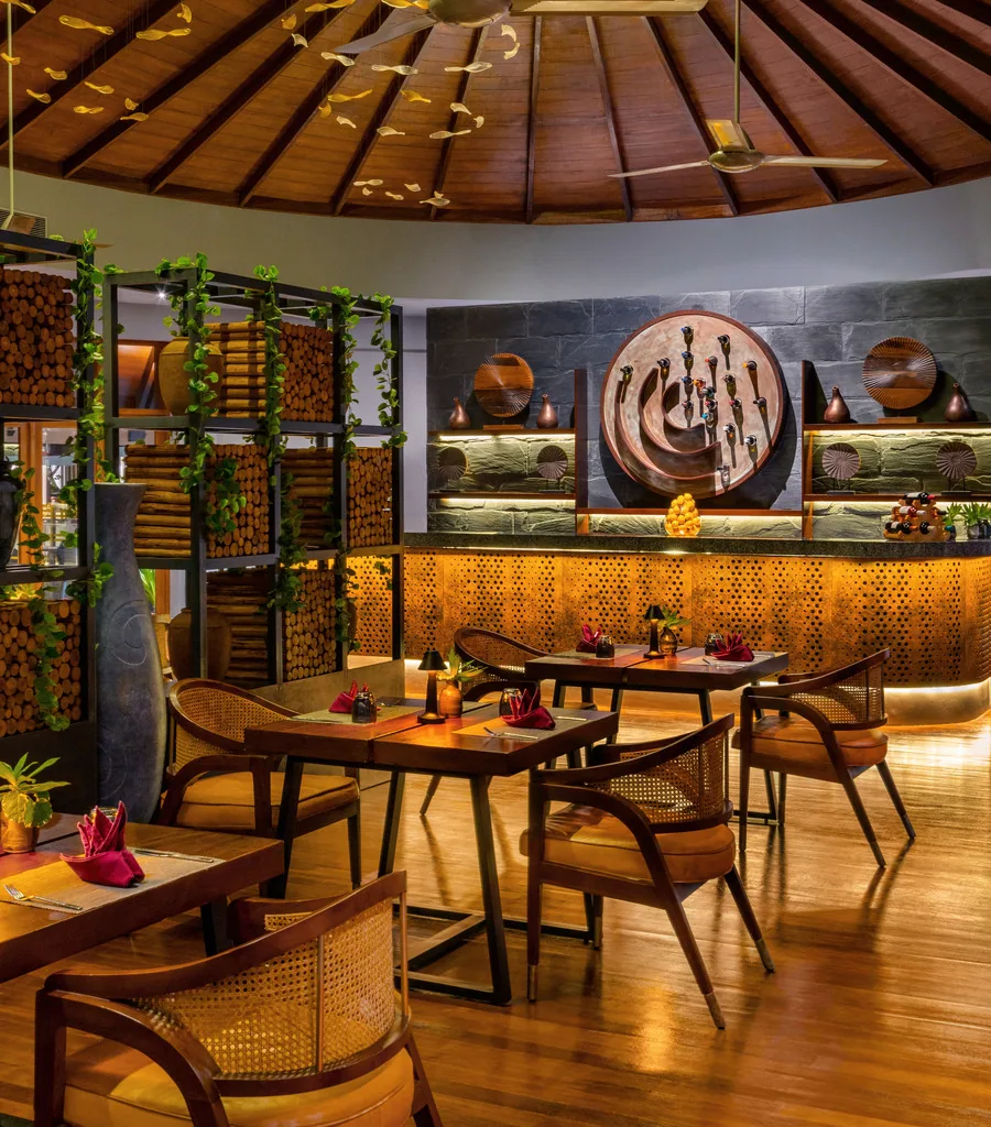 Bandos Maldives, dining, North Malé Atoll, Maldives, Themed restaurant interior: distinctive circular timber radial-rafter ceiling forming a yurt-like vault, a large round wood-plate panel as the wall feature, stacked-log decorative dividers with vine-trail details, rattan-back dining chairs with mustard-cushion seats, warm timber floor under ambient golden lighting.