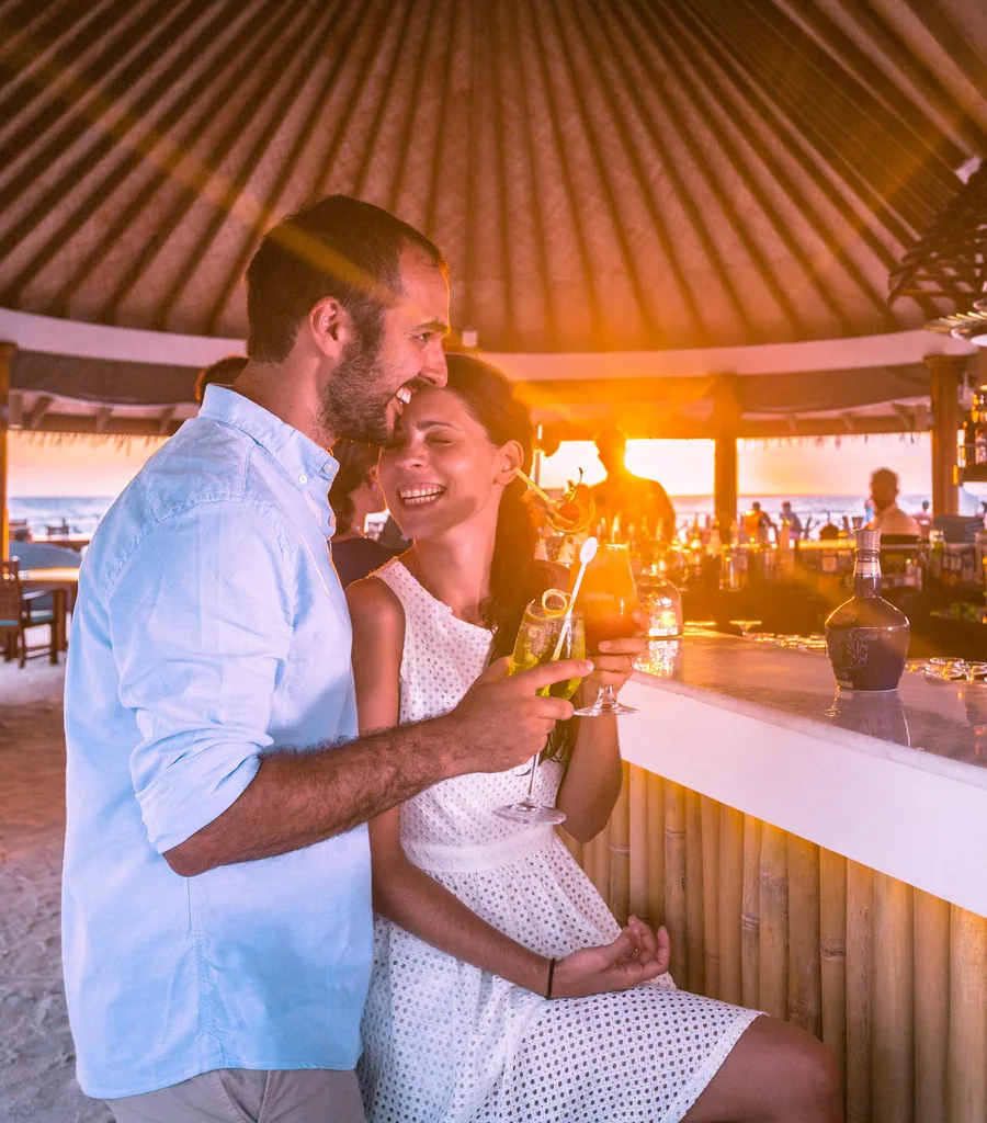 Bandos Maldives, bar, North Malé Atoll, Maldives, Sunset beach bar at the golden hour: a couple toasting with cocktails under the thatched cone-roof bar pavilion (radial palm-frond ceiling), a bamboo-clad bar counter, the sun setting behind them with golden light streaming through, the ocean and white-sand beach visible behind, the property's casual sundown bar setting.