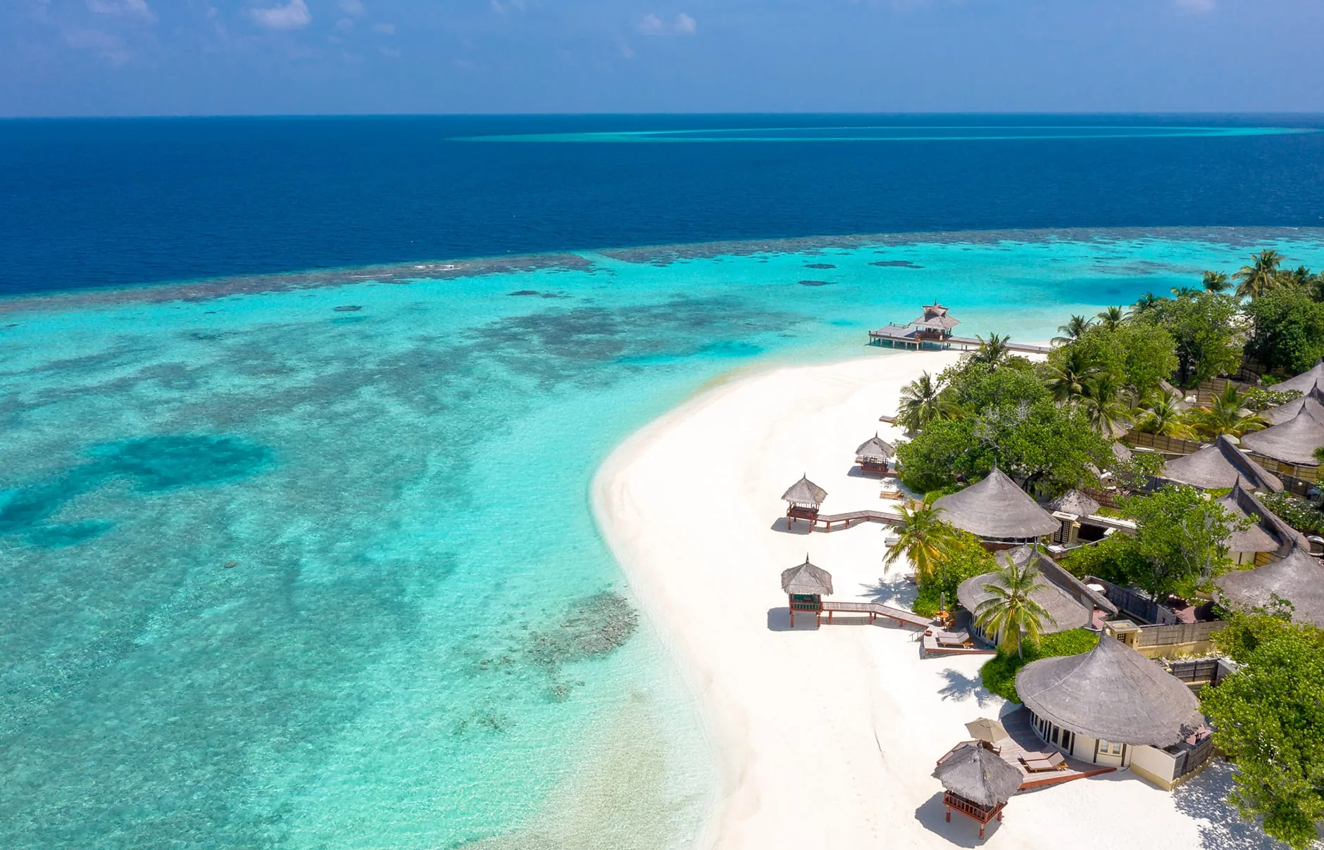Banyan Tree Vabbinfaru, aerial, North Malé Atoll, Maldives, Angled aerial of the western tip, the conical thatched-roof beach villas lined along the white-sand crescent under palm canopy, jetty extending right onto the reef-edge shallow shelf with the spa pavilion at the lagoon end.
