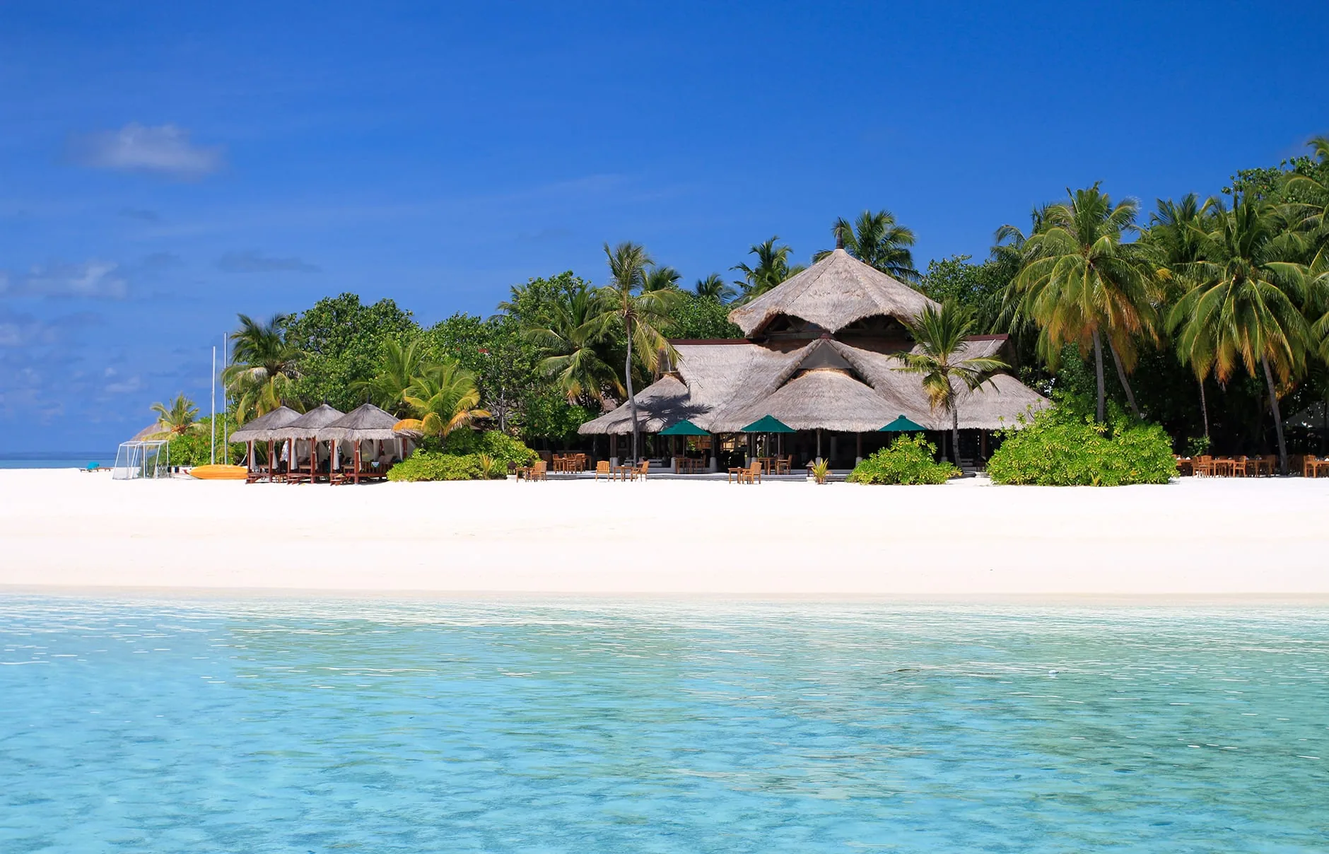 Banyan Tree Vabbinfaru, dining, North Malé Atoll, Maldives, Cluster of thatched conical-roof dining pavilions on the central beach, palm canopy framing the structure, the lagoon foreground reading clear turquoise across the reef shelf.