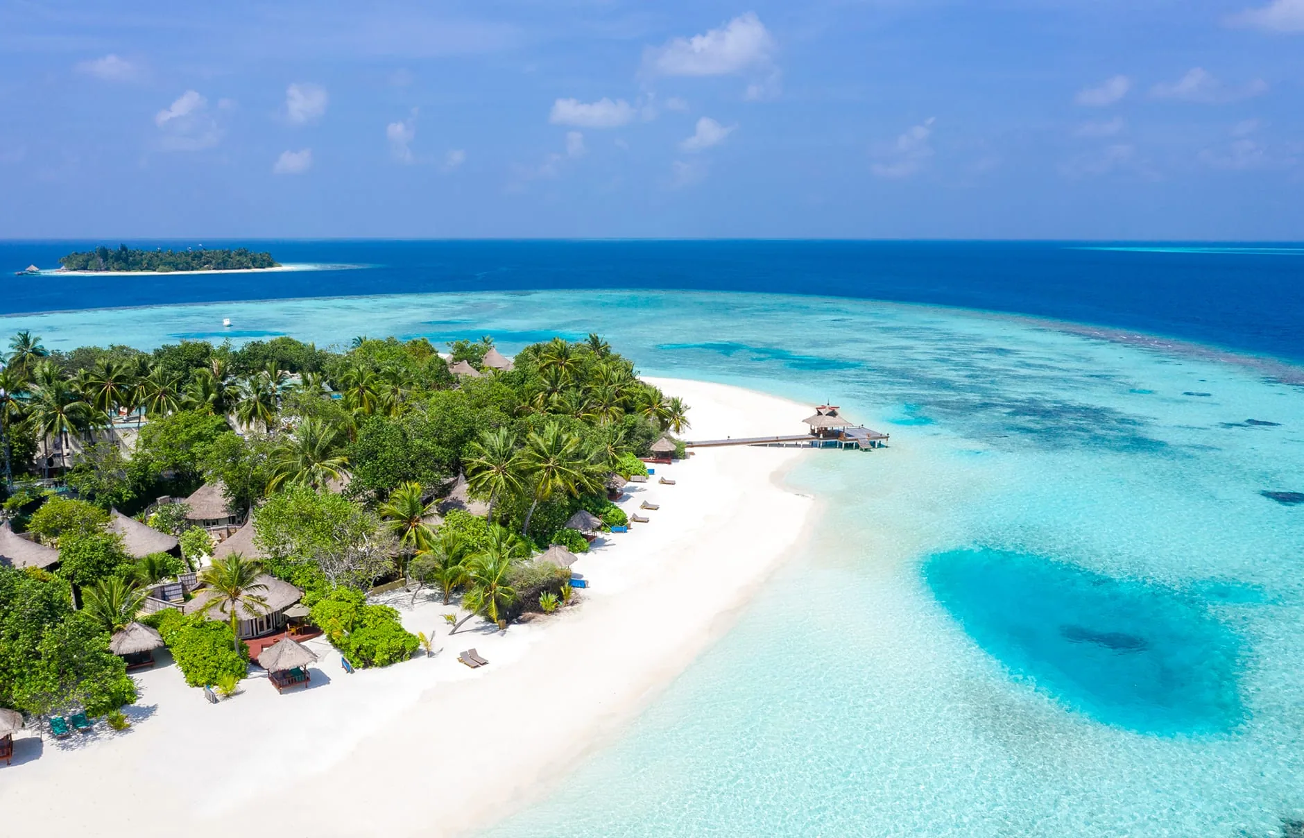 Banyan Tree Vabbinfaru, setting, North Malé Atoll, Maldives, Wide aerial of the western sandbar tip, the white-sand crescent curving into the deeper-blue lagoon shelf on the right, palm-canopy villas tucked into the trees on the left, jetty and spa pavilion extending into the reef edge with the distant atoll islet visible on the horizon.