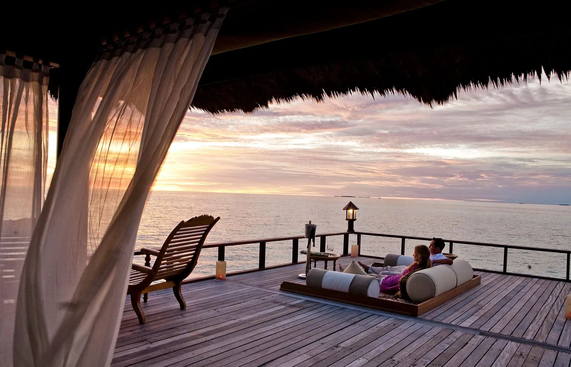 Banyan Tree Vabbinfaru, setting, North Malé Atoll, Maldives, Sunset from an overwater pavilion, gauze curtains framing the view, a couple seated on a padded daybed looking toward the lit horizon lamp, the pastel sky reflecting on the still lagoon and the thatched eaves overhead in silhouette.