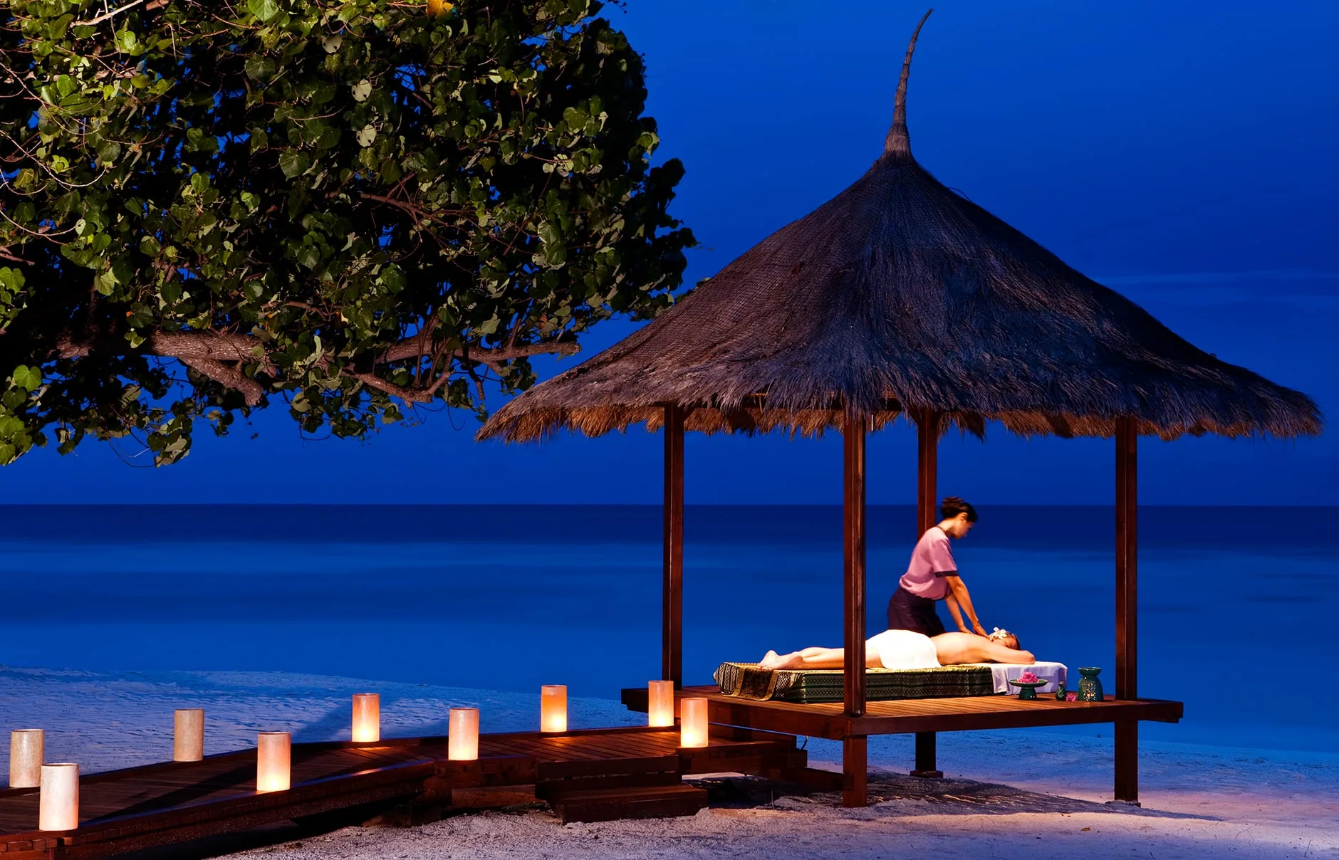 Banyan Tree Vabbinfaru, spa, North Malé Atoll, Maldives, Beach-side spa pavilion at dusk, thatched conical-roof platform with the therapist working on a single massage bed, candle hurricane lanterns lining the timber boardwalk approach, deep-blue dusk sky over the lagoon behind.