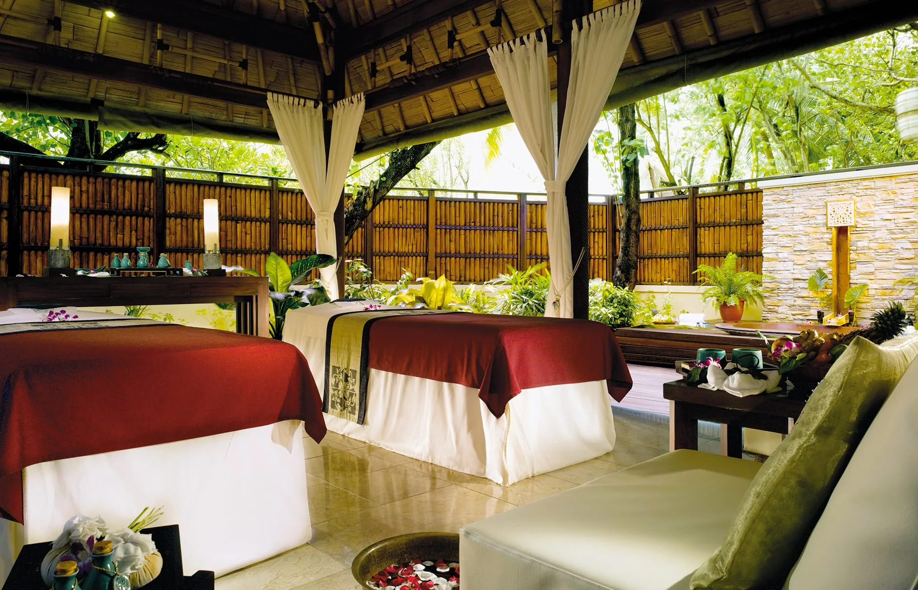 Banyan Tree Vabbinfaru, spa, North Malé Atoll, Maldives, Banyan Tree Spa twin treatment pavilion, two side-by-side massage beds dressed in deep-red linen with white drape, candle lamps on side tables, gauze curtain canopy above, bamboo screen wall and tropical foliage backdrop, the country's first international spa operation since 1995.