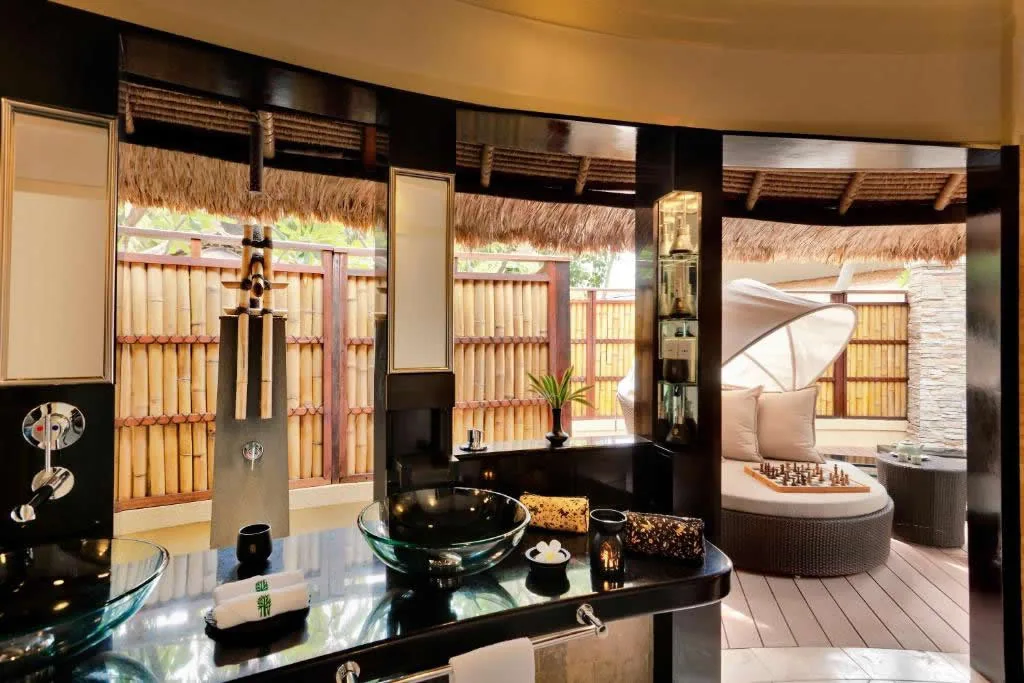 Banyan Tree Vabbinfaru, villa beach, North Malé Atoll, Maldives, Beachfront villa bathroom, signature Banyan Tree Asian-craft detailing: glass vessel basins on a black lacquer counter, dark-timber framing, vertical bamboo-screen wall, view through to the bamboo-and-thatch courtyard daybed with a chess set staged in front of the canopy.