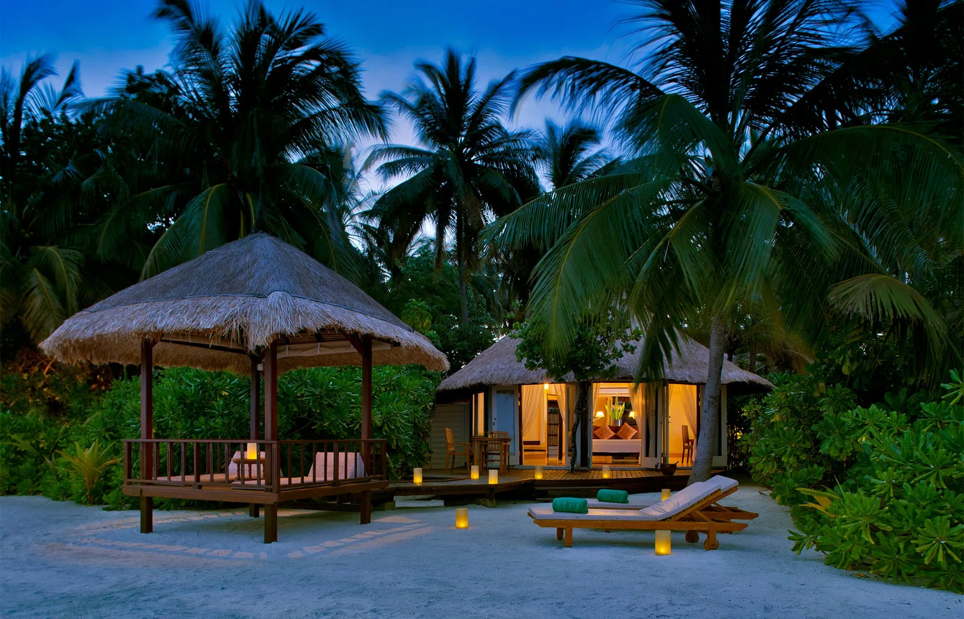 Banyan Tree Vabbinfaru, villa beach, North Malé Atoll, Maldives, Beachfront villa at dusk, thatched conical-roof beach gazebo with hanging-seat daybed, twin sun loungers between candle hurricane lanterns, the villa entry warmly lit through palm trunks behind.