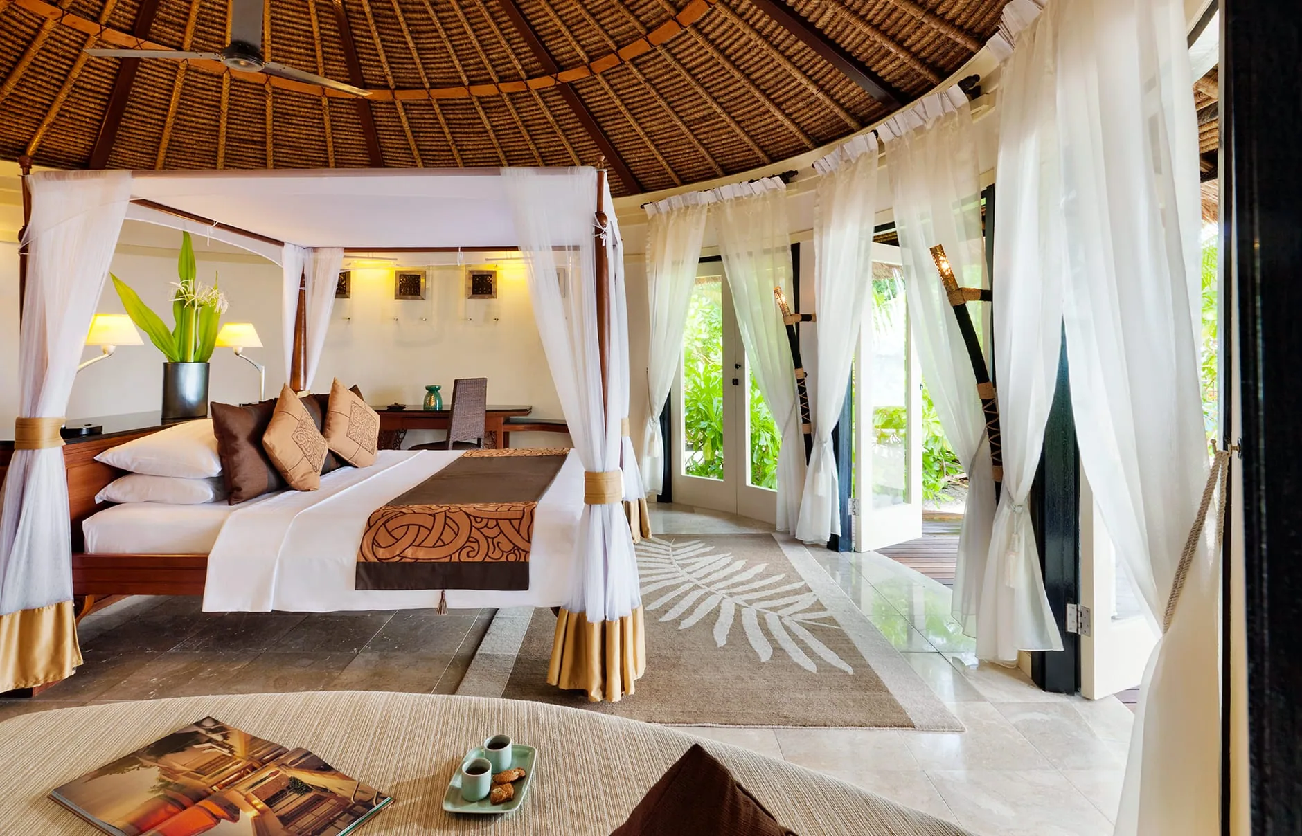 Banyan Tree Vabbinfaru, villa beach, North Malé Atoll, Maldives, Beachfront Pool Villa bedroom interior, signature Balinese round thatched conical ceiling with woven bamboo lining, dark-timber four-poster bed with mosquito-net canopy, copper-and-brown patterned bed runner, gauze curtains open onto the timber deck, marble floor with palm-frond inlay rug.