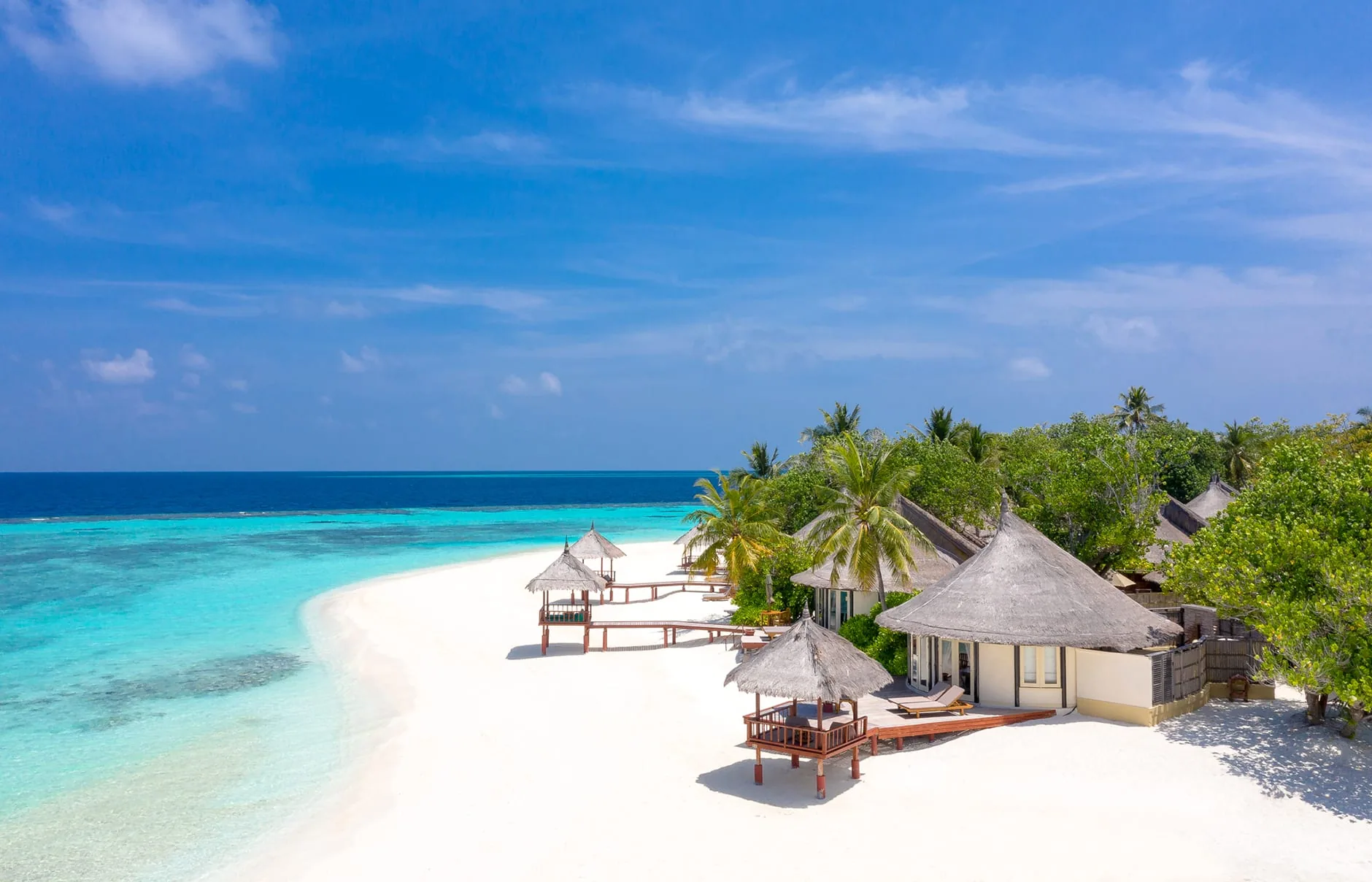 Banyan Tree Vabbinfaru, villa beach, North Malé Atoll, Maldives, Beachfront villa row from a lateral drone perspective, the rhythm of conical thatched-roof villas spaced along the crescent with private timber decks and beach gazebos opening directly onto the sand.