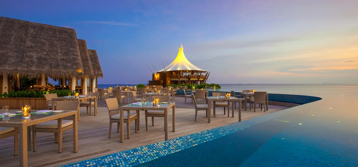 Baros Maldives, dining, North Malé Atoll, Maldives, The Lime Restaurant: beachside open-deck dining at twilight with rattan dining chairs at multiple tables, candle lanterns on each table, the mosaic-tile pool edge in the foreground, the iconic Lighthouse Restaurant white-sail pavilion visible across the lagoon in the mid-distance, pastel peach-orange sunset sky over the calm horizon.