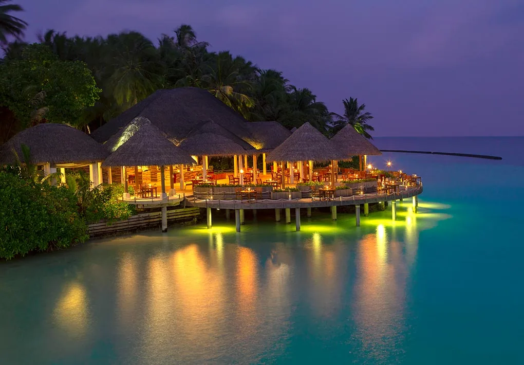 Baros Maldives, dining, North Malé Atoll, Maldives, Cayenne Grill at twilight: the over-water cluster of thatched-cone roof open-air pavilions on stilts above the calm lagoon, warm amber lights glowing through the open structure, dramatic light reflections shimmering on the purple twilight water, dense palm canopy framing the back, the grill house and dry-aged-steak concept captured in property-signature lighting.