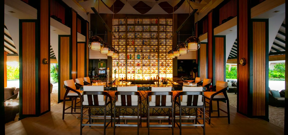 Baros Maldives, bar, North Malé Atoll, Maldives, Sails Bar interior: a striking floor-to-ceiling backlit bar back wall with a stacked-liquor-bottle grid display in golden glow, copper-domed pendant lights, blonde-wood bar stools with orange cushions, dark walnut bar counter, vertical orange-and-walnut wall paneling, a white-and-orange striped tent ceiling structure visible at the top.