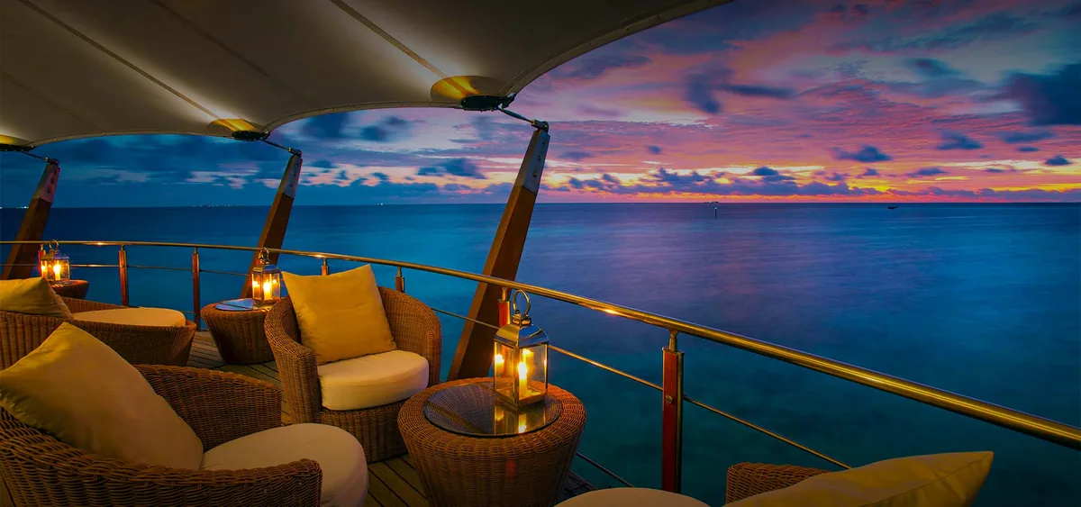 Baros Maldives, bar, North Malé Atoll, Maldives, The Lighthouse Lounge: the upper-floor cocktail lounge of the Lighthouse Restaurant building, woven-rattan armchairs paired with yellow-and-cream cushions, candle lanterns on rattan side tables, the white tensile-canvas roof curve visible at the top edge, brass-and-wood railing, a dramatic peach-purple-blue sunset sky over the calm ocean horizon.