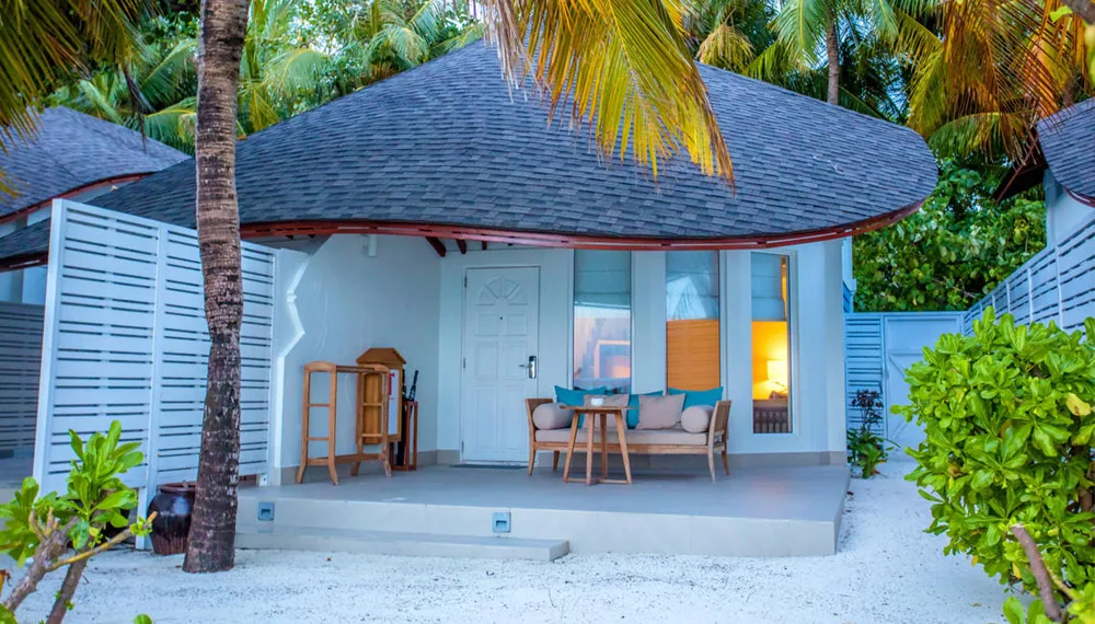 Centara Grand Island Resort & Spa Maldives, villa beach, Alif Dhaal (South Ari) Atoll, Maldives, Beach Suite exterior, shingle-tile umbrella-shape roof above white walls with the turquoise-blue door and matching pillows on the porch bench, white woven privacy panels framing the entry.