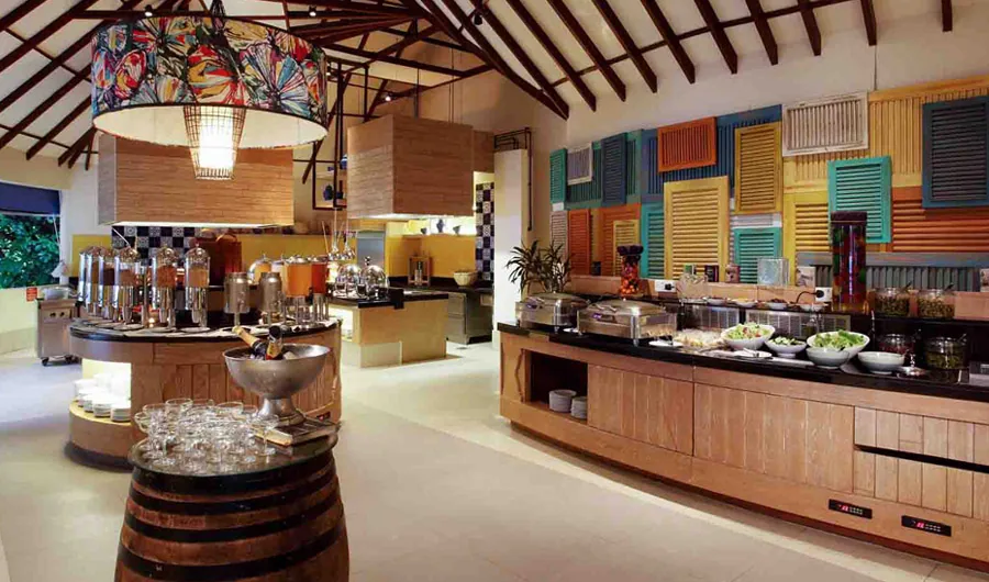 Centara Grand Island Resort & Spa Maldives, dining, Alif Dhaal (South Ari) Atoll, Maldives, Reef main buffet interior with the timber-rafter ceiling, a floral fabric pendant chandelier overhead, the multi-color louvered-shutter feature wall (orange, blue, green and teal panels) anchoring the buffet line, wine-barrel and dispenser station at the foreground.