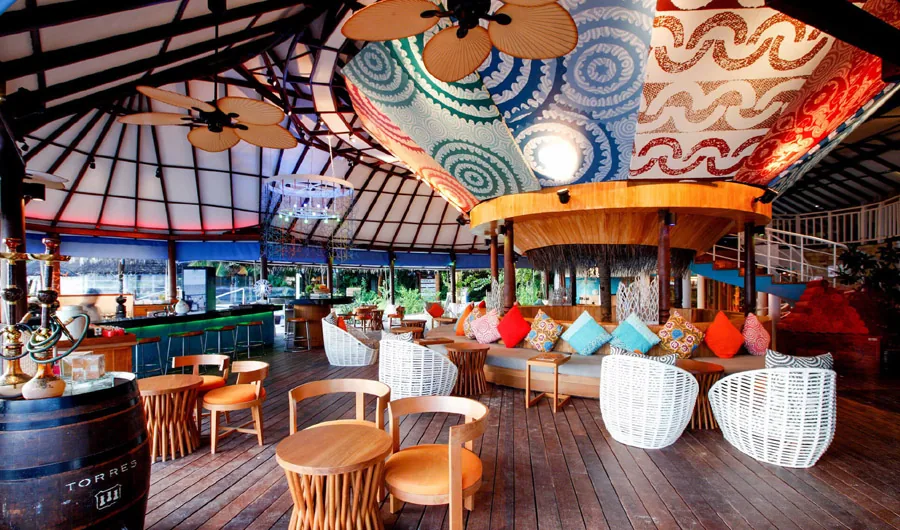 Centara Grand Island Resort & Spa Maldives, dining, Alif Dhaal (South Ari) Atoll, Maldives, Coral Bar and Lounge interior, peaked-roof pavilion with the signature hand-painted canopy underside (blue, orange and yellow circular wave-pattern motifs), white-rope egg-style lounge chairs, the bar with colorful cushion banquette and dramatic floral pendants.