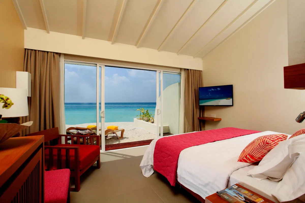 Centara Ras Fushi Resort & Spa, villa beach, North Malé Atoll, Maldives, Beach Villa interior: peaked-vault ceiling with timber beams, king bed with white linens and red velvet runner with red striped accent cushions, sliding glass doors revealing the private beach terrace with sun loungers (yellow towels) and the brilliant turquoise lagoon, the red-cushioned timber armchair in the corner.