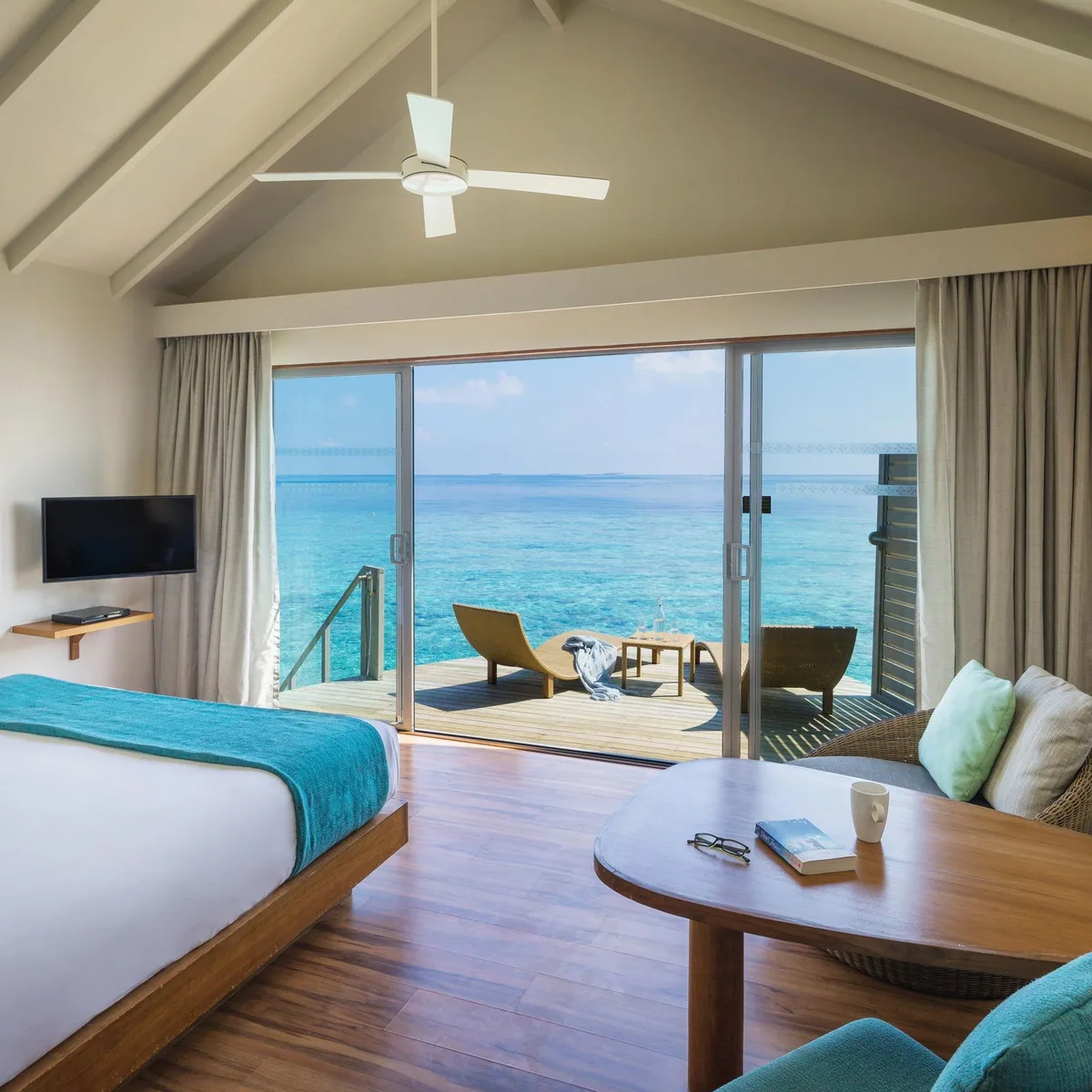 Centara Ras Fushi Resort & Spa, villa overwater, North Malé Atoll, Maldives, Over-water Villa interior: peaked white-vaulted ceiling with a contemporary ceiling fan, king bed with teal accent runner, sliding glass doors revealing the private deck with twin chaise loungers and a small side table over the lagoon, settee with green cushions in the foreground, round timber coffee table, warm walnut floor.