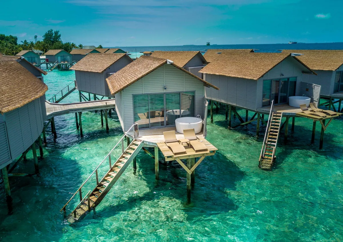 Centara Ras Fushi Resort & Spa, villa overwater, North Malé Atoll, Maldives, Deluxe Spa Over Water Villa exterior: white-walled thatched-roof villas on stilts with the signature circular Swirl Pool jacuzzi feature on the private deck, twin sun loungers, descending timber stairs to direct lagoon access, the crystal-turquoise water with reef visible beneath, the neighbouring villas in the row.