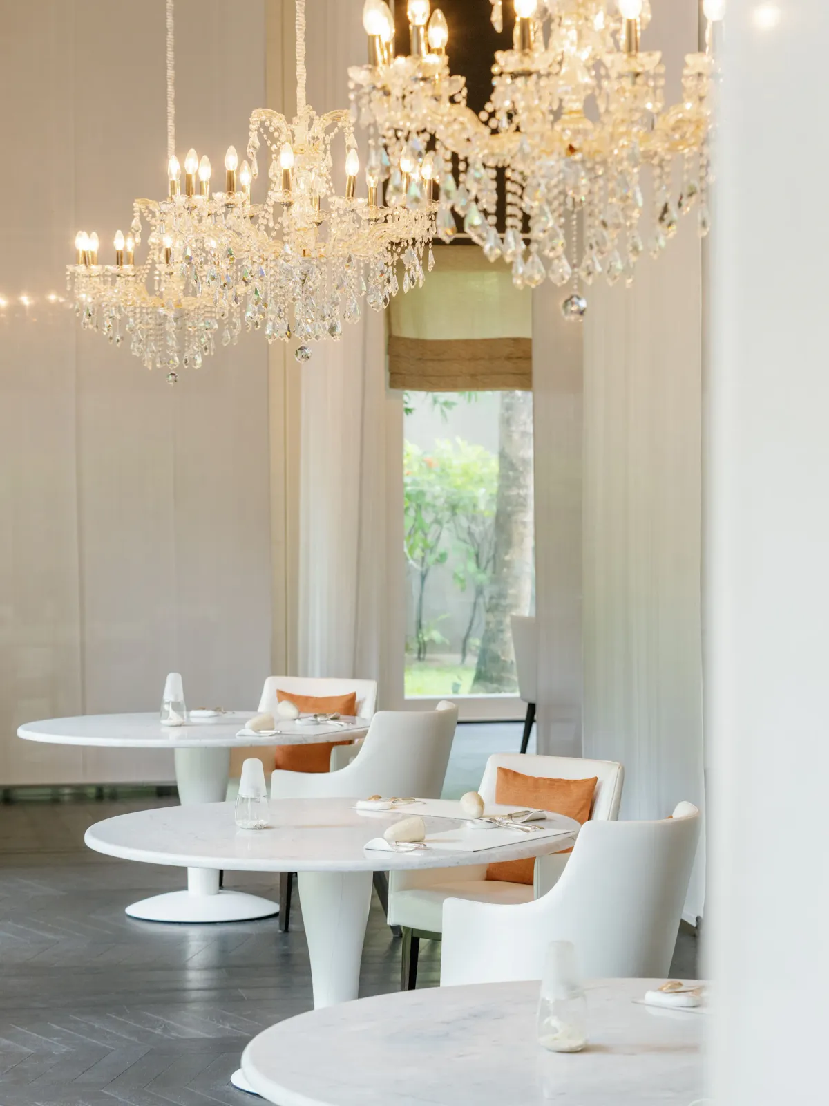 Le 1947 dining room interior with twin crystal chandeliers, white round marble tables and orange-cushion white chairs at Cheval Blanc Randheli.