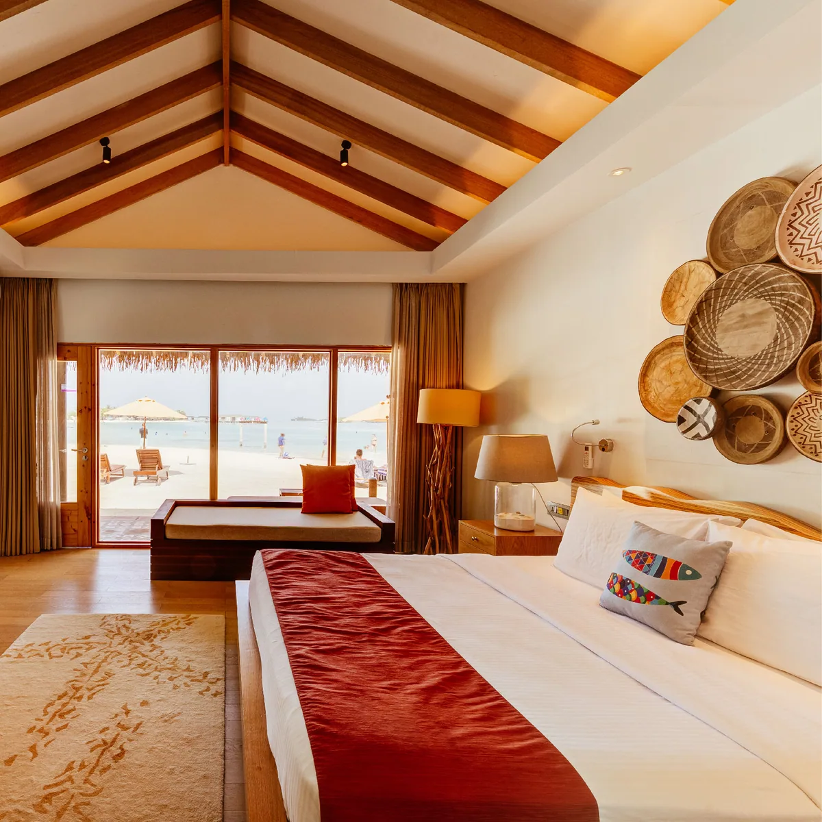 Cinnamon Dhonveli Maldives, villa beach, North Malé Atoll, Maldives, Beach Villa interior detail: vaulted timber-rafter ceiling, king bed with white linens and red velvet runner accent, the signature collection of five woven baskets in varied geometric patterns mounted above the headboard as wall art, terracotta-tile floor, sliding glass doors revealing the beach with sun parasols and the ocean horizon.