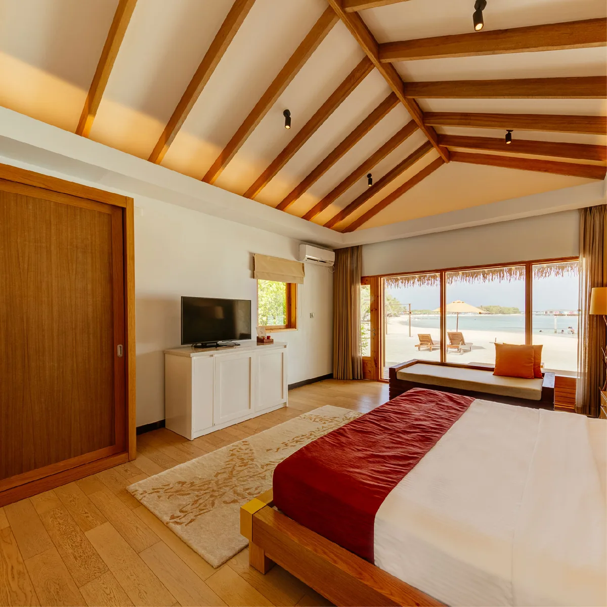 Cinnamon Dhonveli Maldives, villa beach, North Malé Atoll, Maldives, Beach Villa wider spatial layout: vaulted ceiling with exposed timber-beam structure, light parquet floor, TV stand at the entrance side, king bed with red runner at the right edge, large sliding glass doors revealing beach sun loungers with orange parasol and the ocean horizon beyond, the full villa interior layout in single frame.