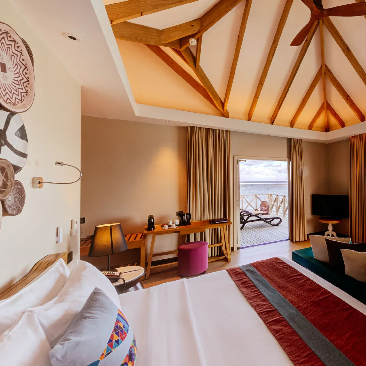 Cinnamon Dhonveli Maldives, villa overwater, North Malé Atoll, Maldives, Water Bungalow tribal-decor interior: vaulted timber-beam ceiling with ceiling fan, king bed with red-and-grey striped runner, signature wall art panel featuring large round wood plates painted with tribal-pattern geometric motifs, pink ottoman accent, sliding glass doors revealing the over-water deck with a sun lounger and the ocean view.