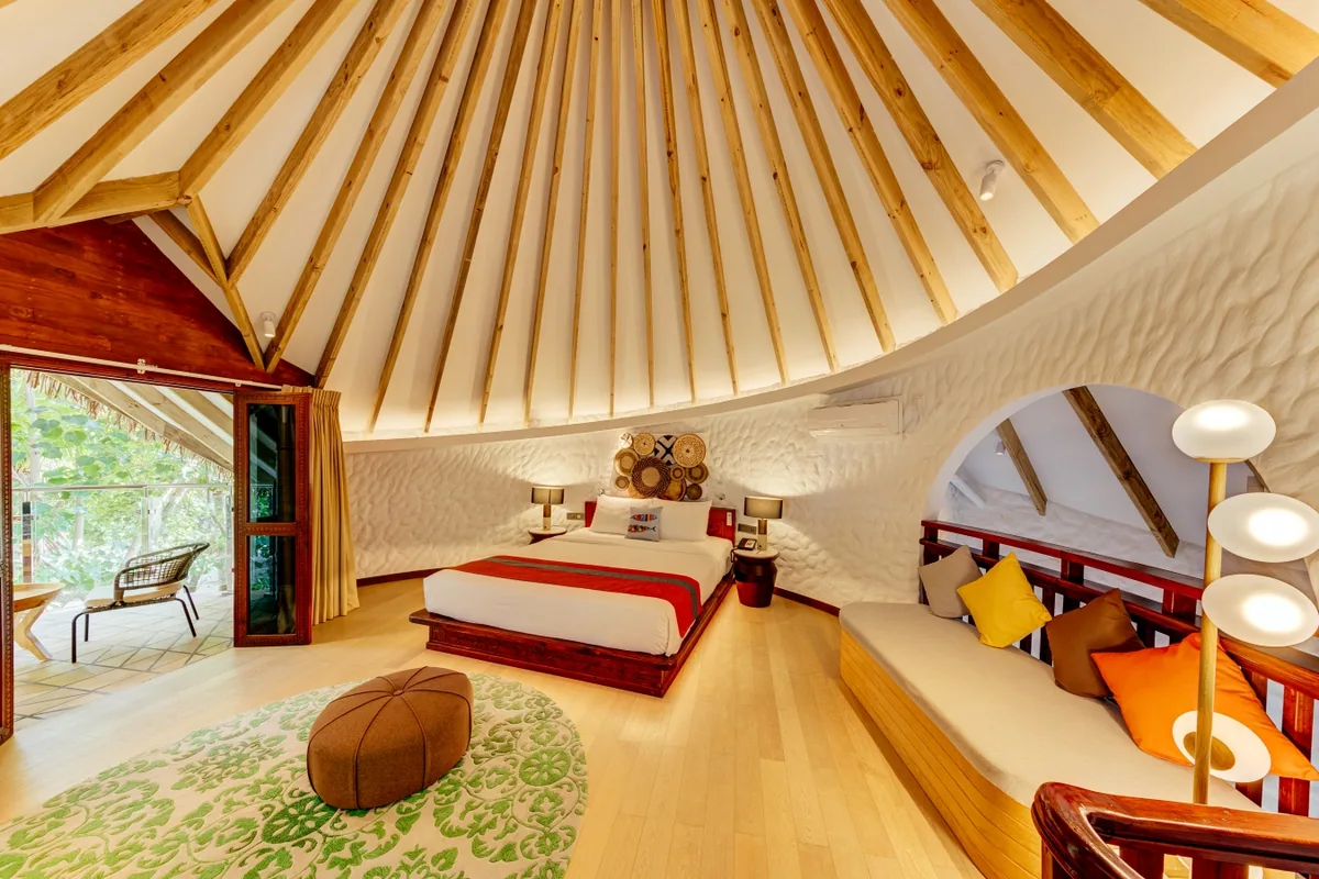 Cinnamon Dhonveli Maldives, villa suite, North Malé Atoll, Maldives, Beach Suite with Pool interior: distinctive dome-shaped ceiling with radial timber rafters (signature property architecture forming a yurt-like vault), white textured-stucco wall finish, king bed against the curved wall with a small basket wall accent, a half-moon green-print rug on the light parquet floor, banquette seating with multi-colour cushions at the right edge, oval mirror, sliding door to the outdoor courtyard.