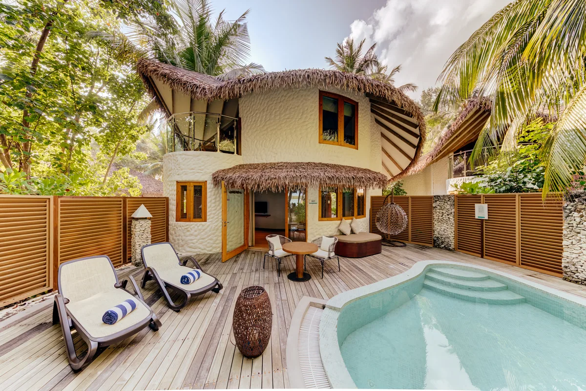 Cinnamon Dhonveli Maldives, villa suite, North Malé Atoll, Maldives, Beach Suite with Pool exterior: distinctive curved-organic stucco architecture with two-tier peaked thatched roof (the property's signature shell-form villa exterior), private courtyard with a kidney-shape plunge pool, twin sun loungers on the timber deck, a sofa daybed plus a suspended egg chair, slatted timber privacy fence, tropical foliage enclosing the courtyard.