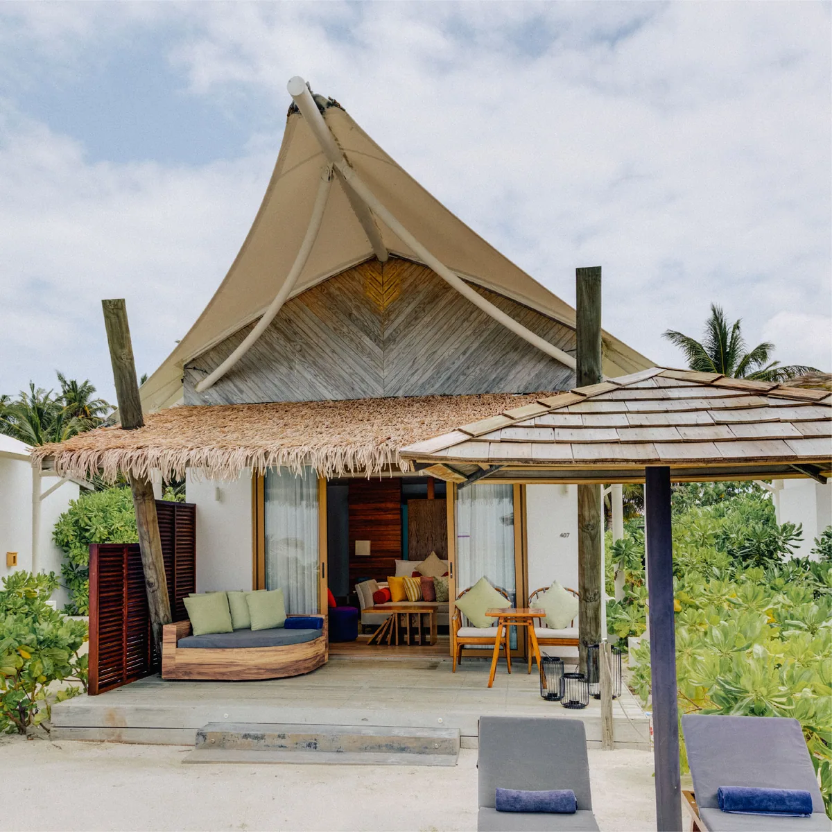Cinnamon Hakuraa Huraa Maldives, villa beach, Meemu Atoll, Maldives, Beach Bungalow exterior: distinctive tent-peak roof (white canvas tensile structure with thatched eaves below), the open-front pavilion shelter housing a crescent-curved daybed with green cushions plus a small lounge seating area, blue-trimmed louvre shutters flanking the door, the side cabana with a separate daybed, sandy palm-tree garden setting.