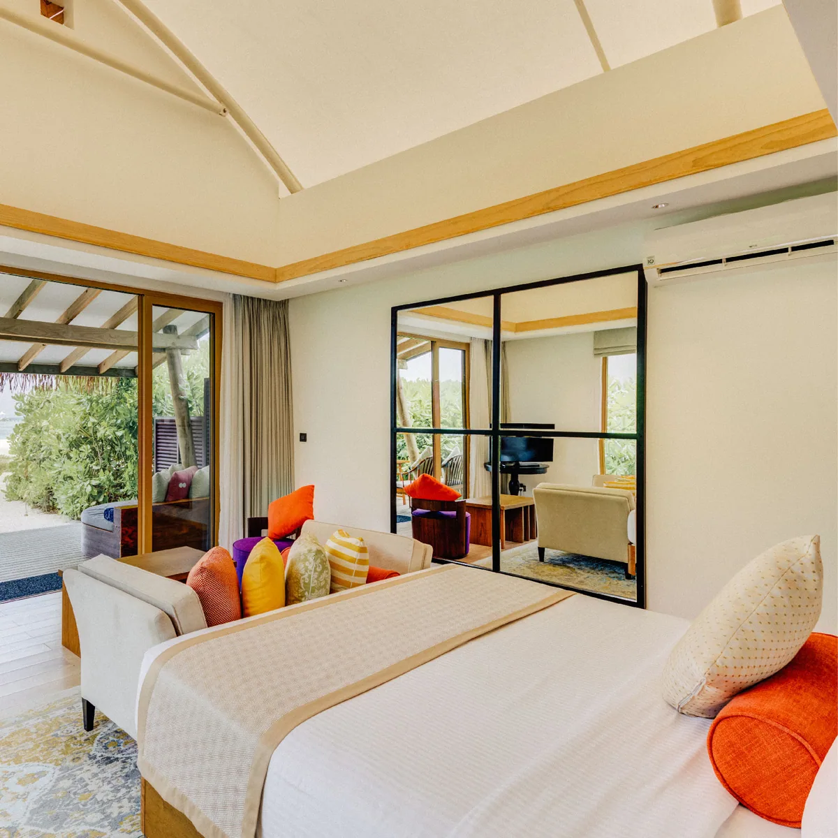 Cinnamon Hakuraa Huraa Maldives, villa beach, Meemu Atoll, Maldives, Beach Bungalow interior: the signature vaulted tent-peak fabric ceiling visible from inside, a king bed with foot-of-bed bench scattered in vibrant orange, yellow, purple, and striped cushions, a large mirrored wall reflecting the room, sliding glass doors revealing the outdoor entrance and small patio.