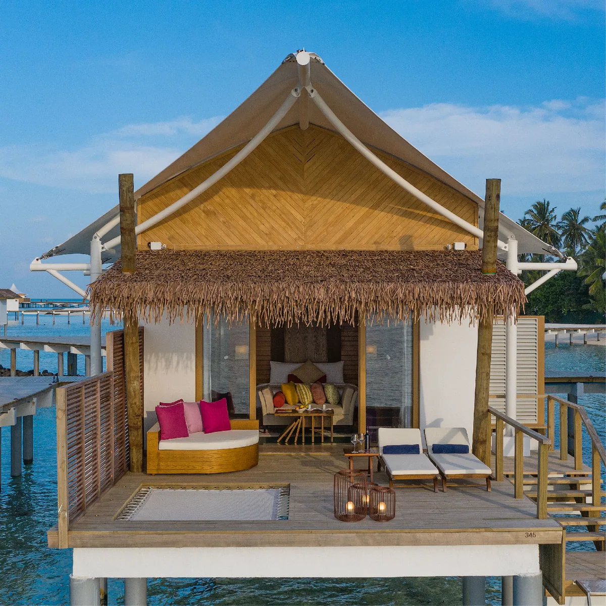 Cinnamon Hakuraa Huraa Maldives, villa overwater, Meemu Atoll, Maldives, Water Bungalow exterior on stilts: the same tent-peak signature roof now suspended over the lagoon, the private deck with a crescent daybed (pink, yellow, and turquoise cushions), twin sun loungers, a small plunge pool with glass-floor showing the lagoon below, basket lanterns on the deck, the connecting jetty visible at the edges.