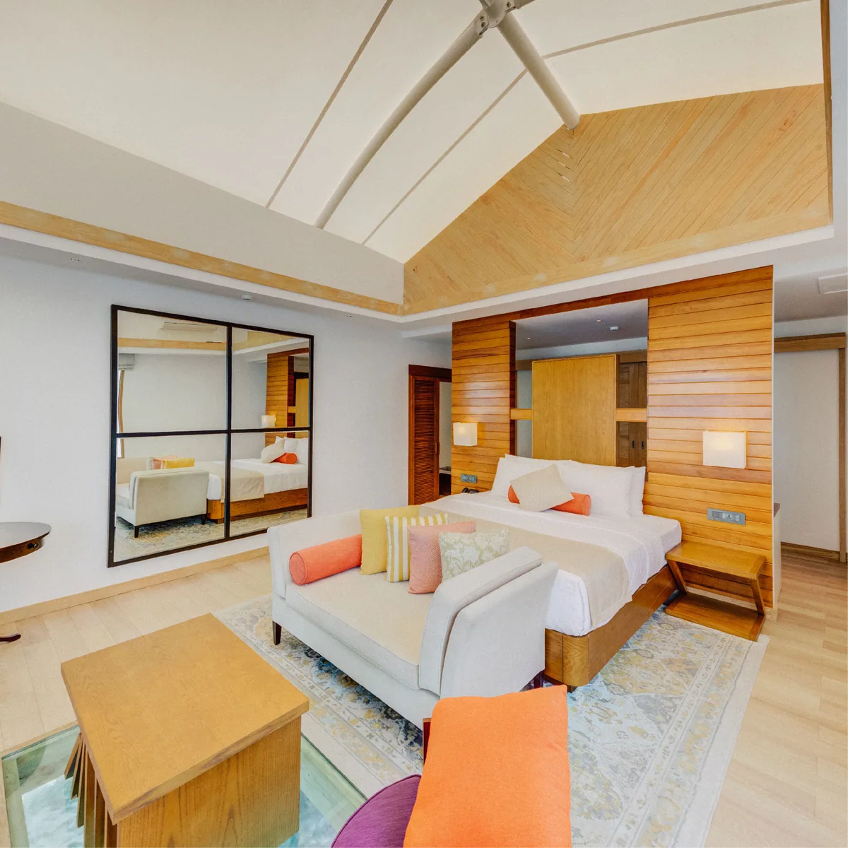 Cinnamon Hakuraa Huraa Maldives, villa overwater, Meemu Atoll, Maldives, Water Bungalow interior: the signature tent ceiling visible from inside, a chevron-pattern timber-clad feature wall behind the king bed, white settee at the foot of the bed with pink, orange, and yellow cushions, ceiling fan, and the signature glass-floor cutout in the foreground revealing the lagoon water beneath the villa.