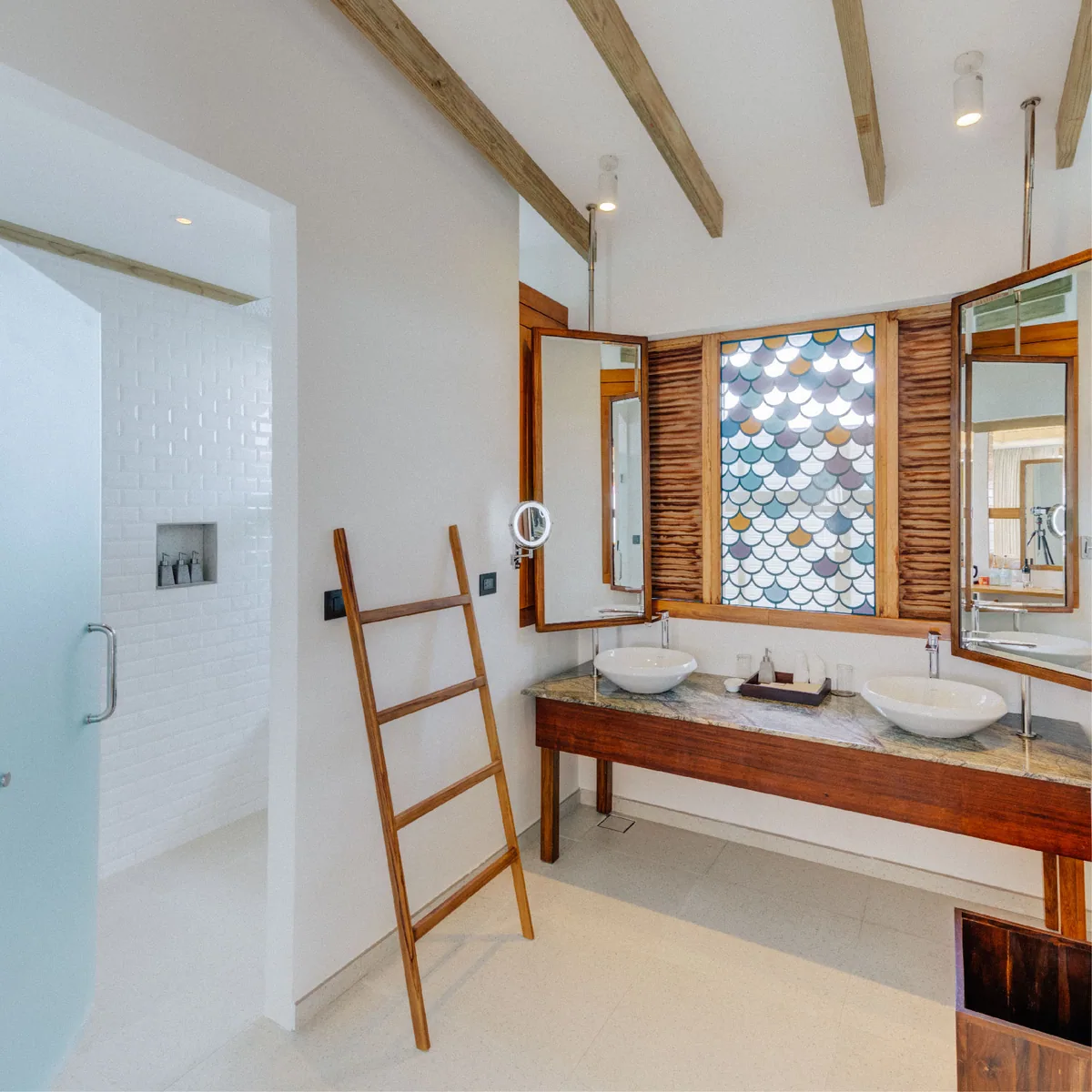 Cinnamon Hakuraa Huraa Maldives, villa bath, Meemu Atoll, Maldives, Water Bungalow bathroom: twin vessel sinks set on a marble-and-timber vanity, a distinctive fish-scale stained-glass window (signature design element with multi-colour scalloped panes), a timber towel ladder, exposed timber roof beams, the walk-in shower with white subway tile visible through the doorway at left.