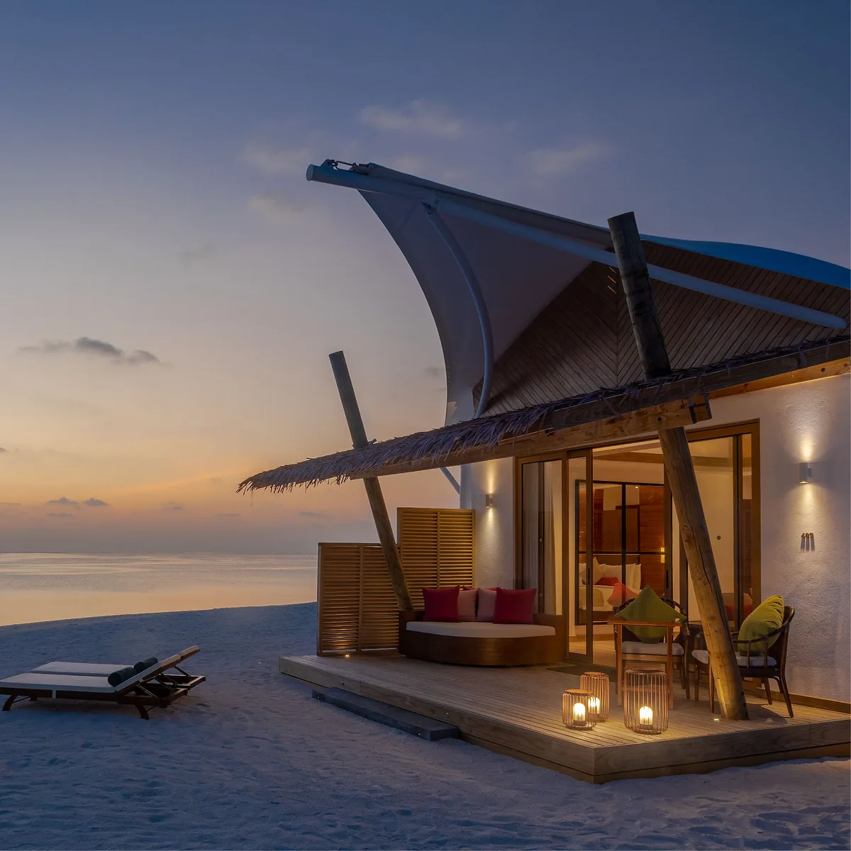 Cinnamon Hakuraa Huraa Maldives, villa beach, Meemu Atoll, Maldives, Platinum Beach Bungalow at twilight: the same tent-peak signature roof over a warm-lit interior visible through the open glass doors, the deck with a crescent daybed (red and pink cushions), pillar candle lanterns on the deck floor, twin sun loungers on the white sand to the side, a pastel sunset sky over the lagoon with the day's footprints in the sand.