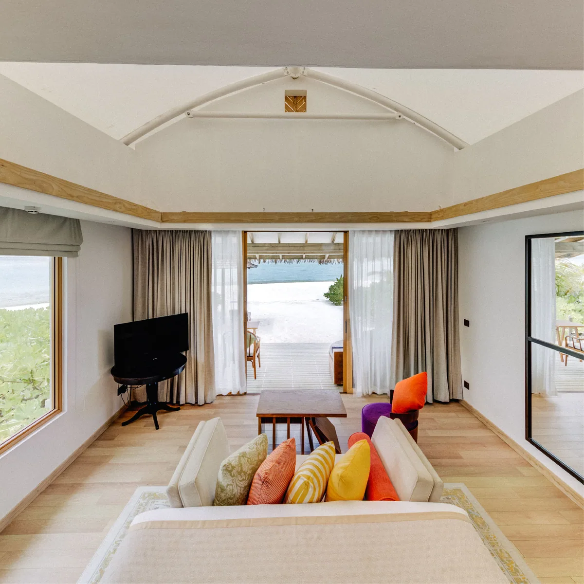 Cinnamon Hakuraa Huraa Maldives, villa beach, Meemu Atoll, Maldives, Platinum Beach Bungalow interior: tent-peak ceiling visible, the wide view looking out through the open glass doors to the beach terrace plus the crescent daybed plus the sun loungers and lagoon beyond, a long settee with colourful cushions in the foreground, gauzy curtains framing the view.