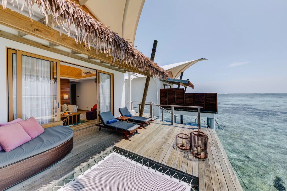 Cinnamon Hakuraa Huraa Maldives, villa overwater, Meemu Atoll, Maldives, Sunset Water Bungalow private deck: the tent-peak roof with thatched eaves visible at the top edge, glass sliding doors revealing the warm interior, a crescent daybed with pink cushions, dual sun loungers with blue cushions, the distinctive over-water hammock-net stretching across the deck edge for relaxing-over-the-water seating, basket lanterns alongside, the divider screen separating this villa's deck from the neighbour.