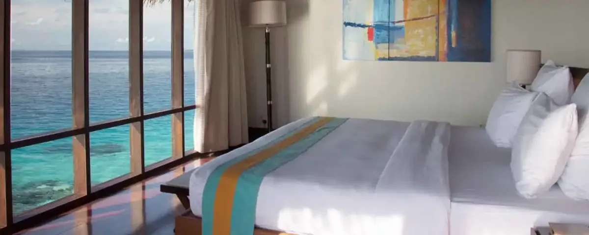Coco Bodu Hithi, villa overwater, North Malé Atoll, Maldives, Water Villa interior: king bed with white linens plus a turquoise-and-orange runner, an abstract blue-orange-teal canvas art panel mounted above the headboard, floor-to-ceiling window revealing the reef-shallow lagoon directly below plus the ocean horizon, gauzy curtains, warm walnut floor.