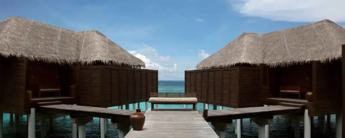 Coco Bodu Hithi, villa overwater, North Malé Atoll, Maldives, Over-water boardwalk view: looking along the access boardwalk between two thatched-roof over-water villas, dark walnut-shutter exterior walls framing the path, a low timber bench at the far end of the boardwalk, terracotta pot accent, the open lagoon and ocean beyond, the property's villa-access circulation captured in single frame.