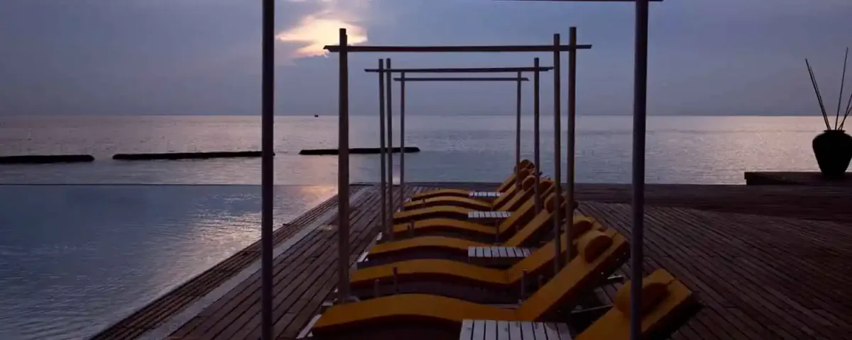 Coco Bodu Hithi, pool, North Malé Atoll, Maldives, Main pool deck at twilight: row of yellow-cushioned sun loungers arranged under a timber-pergola frame structure, the infinity pool edge with breakwater rocks visible in the water just beyond, a dramatic peach-and-purple twilight sky over the calm horizon, the property's signature ambient sundown composition.