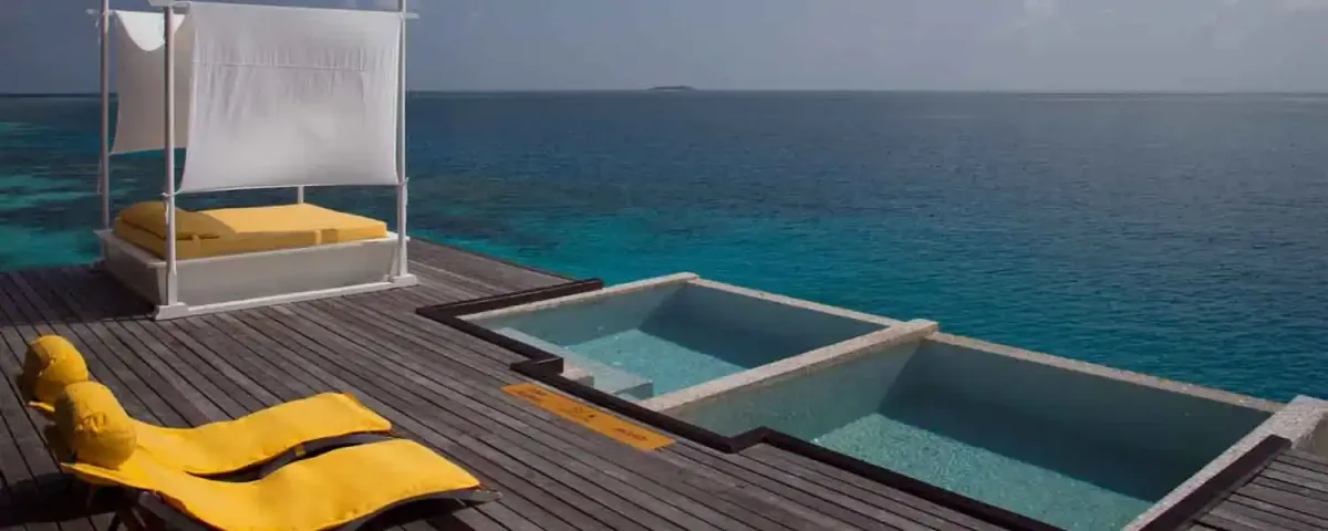 Coco Bodu Hithi, villa overwater, North Malé Atoll, Maldives, Escape Residence private deck: twin yellow-cushioned chaise loungers plus a canopied four-poster daybed with yellow cushions and a white sail-drape, dual-step infinity plunge pools cascading toward the open ocean, the deep cobalt-blue horizon, the property's signature yellow-cushion palette captured across the residence-tier configuration.